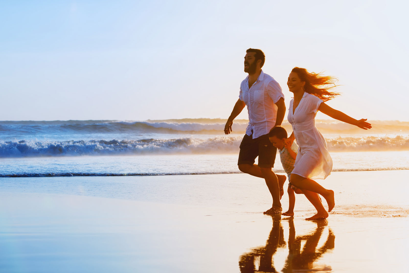 Happy family - father mother baby son hold hands and run together with fun along sunset sea surf on black sand beach. Travel active lifestyle parents with children on tropical summer vacations.